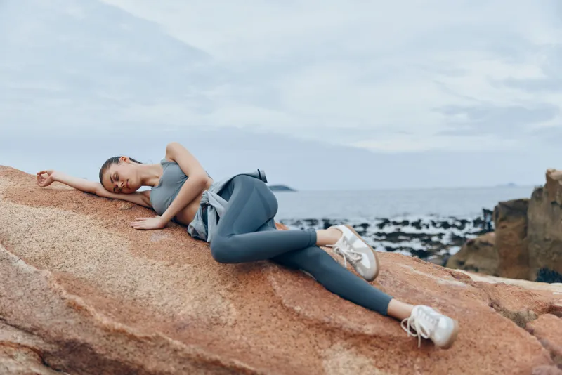 Young woman in sportswear relaxing on a rock by the sea, showcasing a peaceful and relaxed emotion, perfect for healthy lifestyle and fitness concepts