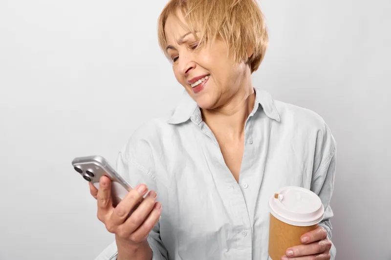 Senior woman holds smartphone, smiles and drinks coffee from a cup, communication and messaging on screen, relaxed casual home studio portrait, white background, closeup