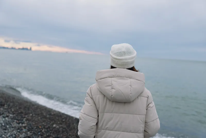 Winter beach scene with a person in a padded hooded coat standing on a pebbled shore, facing the sea under an overcast sky and calm horizon, minimalist and contemplative.