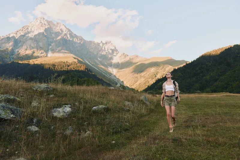 Young woman with long hair walks through a mountain meadow wearing shorts and a white top, enjoying a sunny day in nature with a majestic peak behind her.