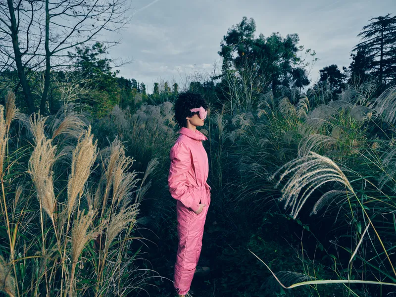 Person in a pink suit stands among tall grasses outdoors, stylish and confident, wearing sunglasses in a natural field setting, highlighting bold fashion.