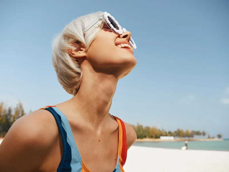 Young woman with short platinum hair smiles up at the sun, wearing sunglasses and a colorful swimsuit on a bright beach, capturing carefree summer energy.