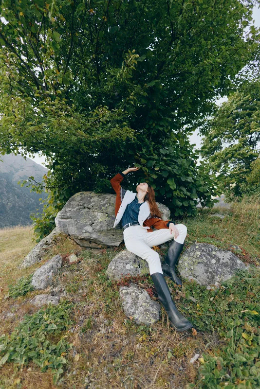 Woman in nature relaxing under a tree on rocks in a countryside meadow, wearing boots and casual outfit, lying back and enjoying peaceful outdoor leisure and scenic landscape.