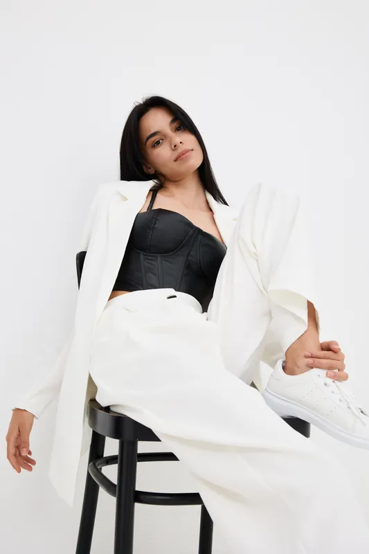 Young woman posing stylishly in white suit and black corset while sitting relaxed on black chair against plain white background, modern fashion and confidence.