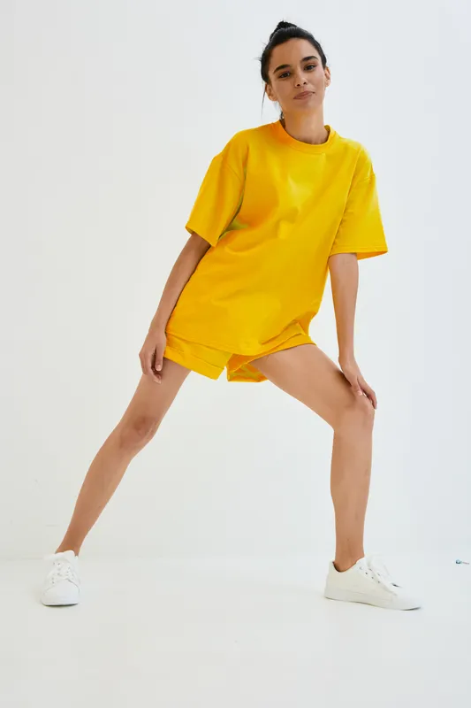 Young woman in bright yellow casual outfit stretching legs in minimalist white studio, demonstrating fitness and healthy lifestyle with sporty white sneakers.