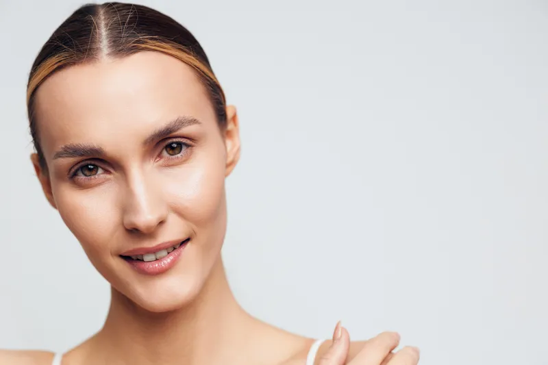 A close-up portrait of a smiling young woman with clear skin and slicked-back hair against a plain white background. The image conveys beauty, confidence, and natural skincare.