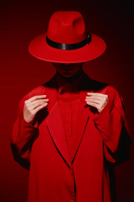 Fashion model wearing matte red hat and coat posing with head down in intense red lighting. Concept of bold style beauty creative fashion statement in studio.