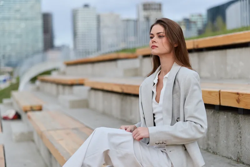 Business woman in light suit sitting outdoors on urban concrete steps with modern cityscape background, looking thoughtful and serious, natural lighting