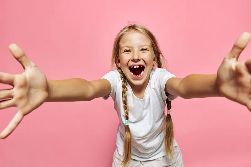 Cheerful smiling girl with braided hair reaching out with open arms on a pink background.