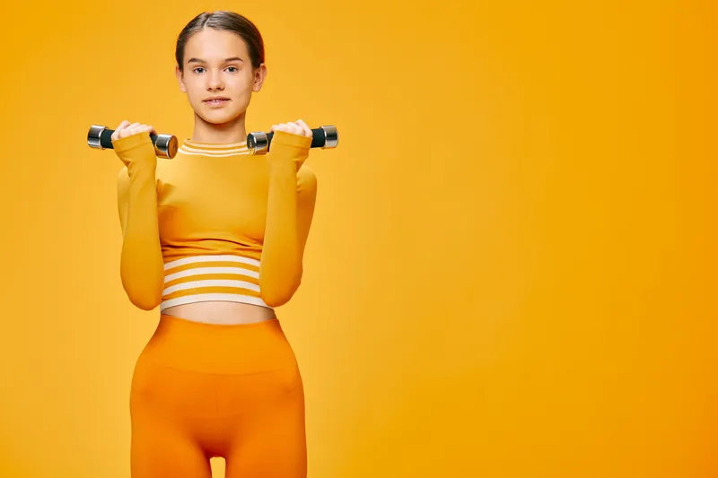Fitness woman in bright orange activewear lifts a dumbbell against a vivid orange background, showcasing strength, focus and athletic energy during a studio workout session.