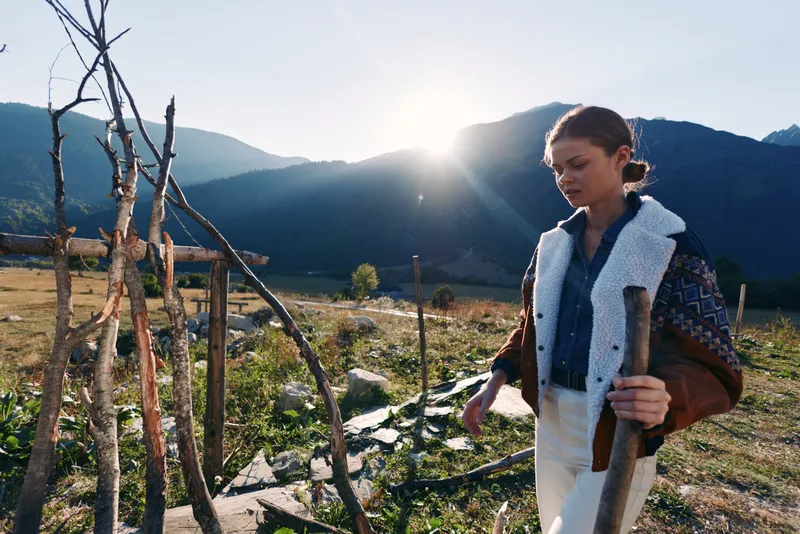 Woman in mountains exploring countryside field at sunset, holding a stick beside a rustic wooden fence. Outdoor portrait with jacket and backlight sunlight over valley scenery.