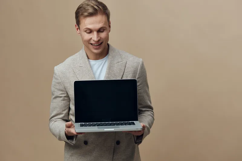 Smiling man holding a laptop in hands wearing stylish beige blazer and white t-shirt, expressing happiness and confidence, standing against plain beige background, studio portrait of cheerful professi