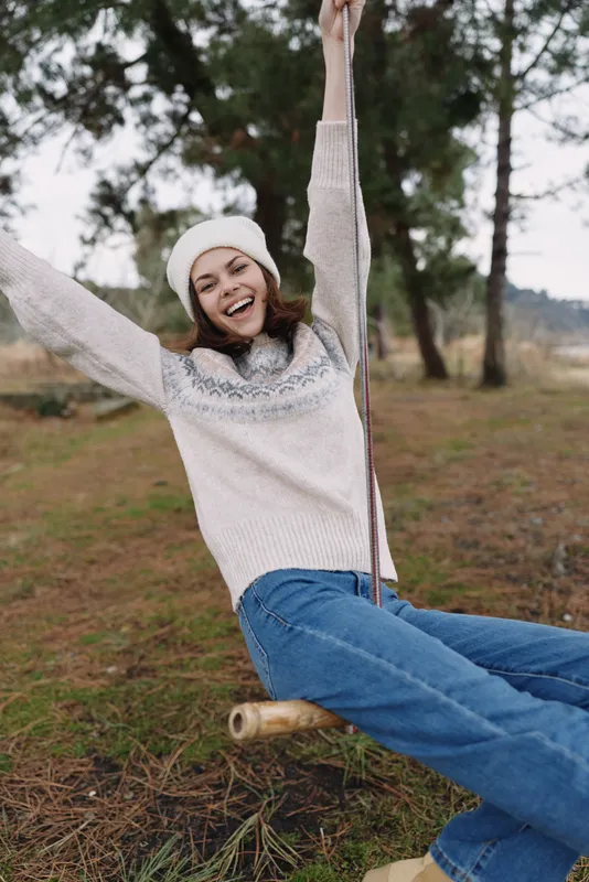 Woman on a wooden swing in a park, wearing a cozy sweater and beanie, smiling with arms up, carefree moment in autumn light among trees and grass, casual outdoor leisure and youthful energy.