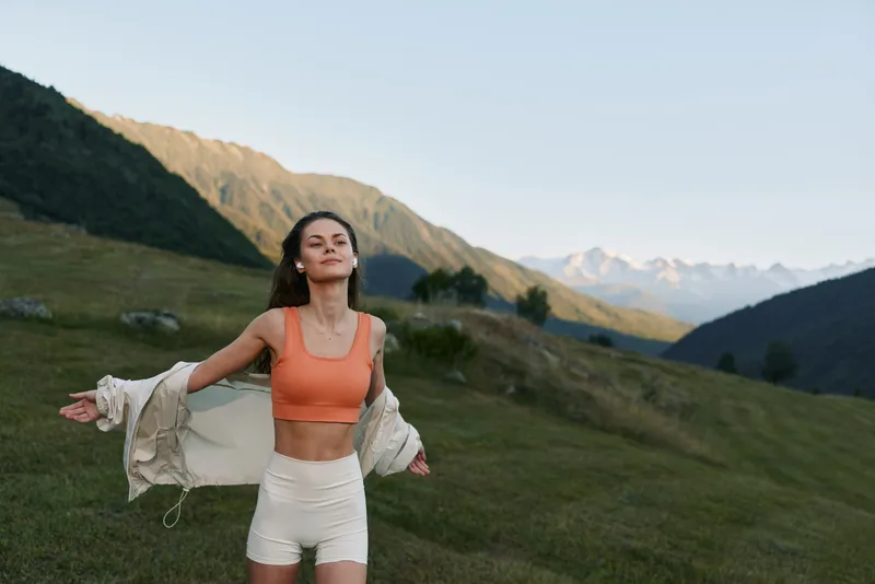 A cheerful woman stands in a scenic meadow with majestic mountains in the background, wearing an orange sporty top and white shorts, expressing freedom and outdoor vitality.