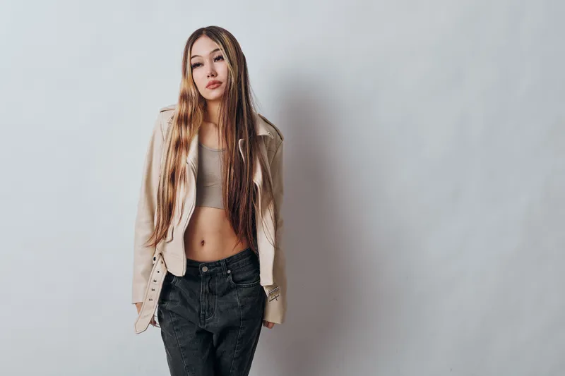 Young woman standing with long hair and casual outfit including beige jacket and black jeans against plain light background with confident posture and expression.