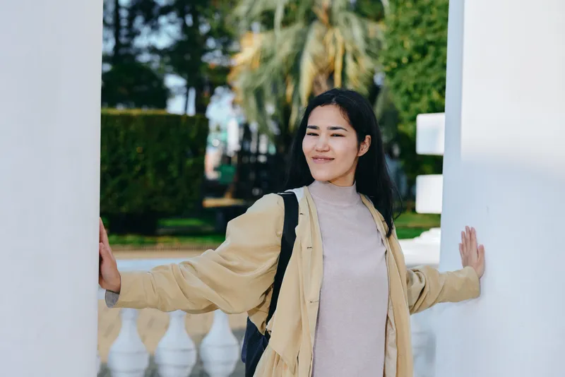 Woman standing at an outdoor doorway, wearing a beige coat, smiling softly, with backpack, in a leafy courtyard, presenting a casual fashion scene and relaxed travel vibe.