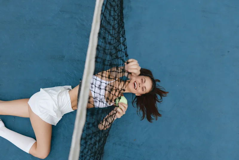 Young female tennis player joyfully lying on the court, holding a tennis ball, wearing sports attire, showcasing athleticism and happiness in a vibrant outdoor setting