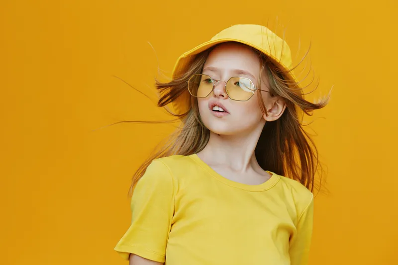 Young girl in a yellow outfit with sunglasses and a cap, posing against a vibrant yellow background, embodying summer vibes and joy