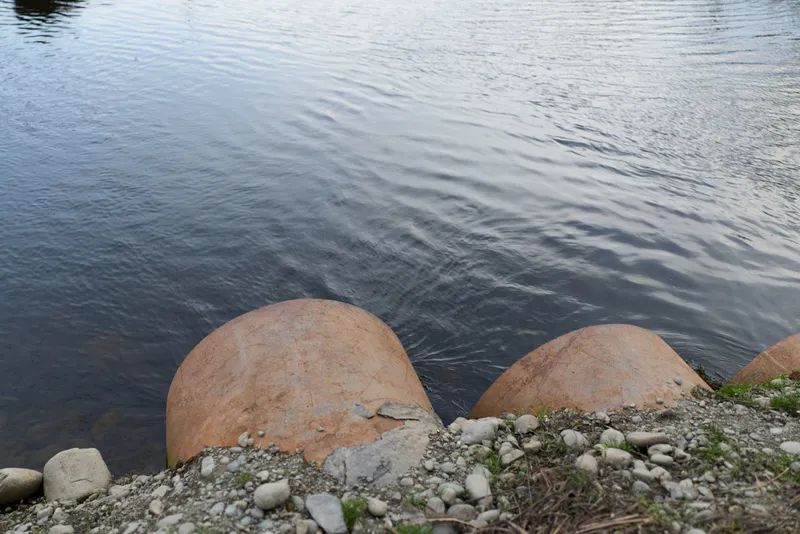 Lake river shoreline scene, water ripples across the surface, rocks and boulders resting on a pebble shore, outdoor calm nature landscape, soft daylight, green moss, tranquil mood