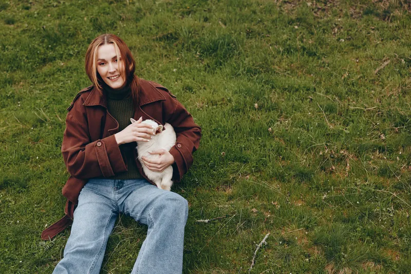 Smiling woman in a brown jacket sits on grass, holding a goat kid. Outdoor scene highlights lifestyle, nature, and affection with a gentle bond between human and animal. Warm, soft lighting creates a
