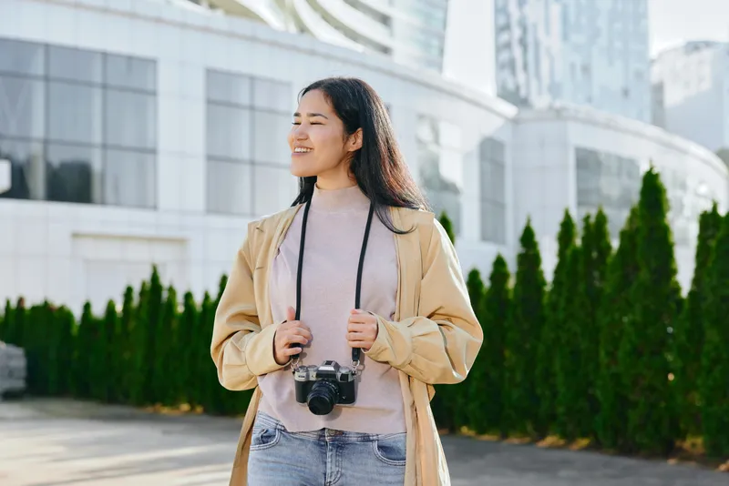 Woman outdoors in an urban setting with camera over her shoulder, smiling, wearing a light trench and knit sweater, modern architecture in background, lifestyle travel and street photography ready