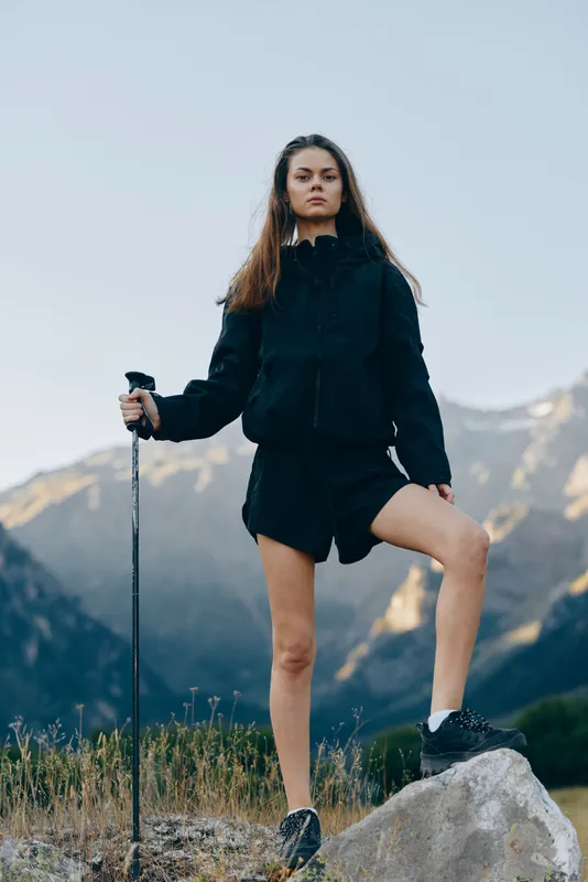 A young female hiker stands on a rocky outcrop in a mountain landscape, gripping a trekking pole. She wears a dark jacket and shorts, looking ahead with calm confidence for the trek.