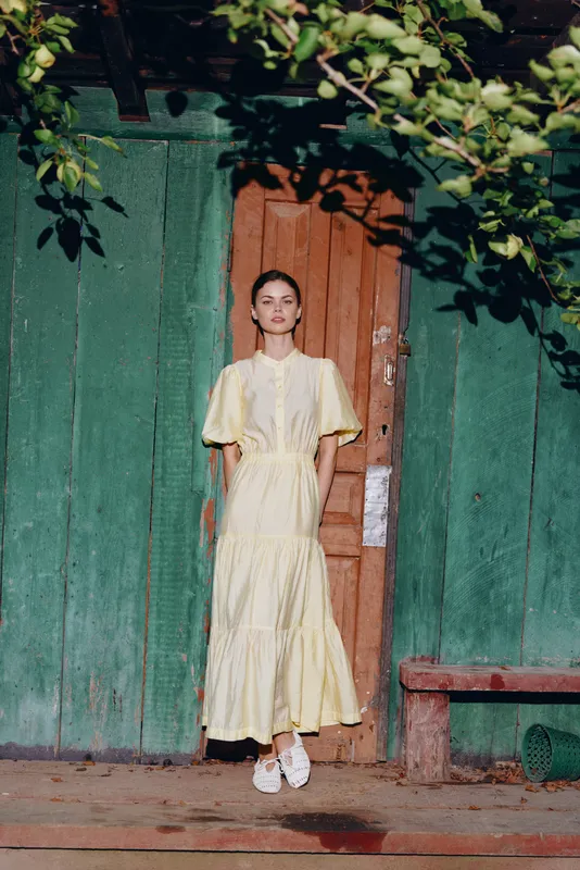 Portrait of a girl wearing a flowing yellow dress standing in front of a green wooden wall and an orange door, exuding calm charm and summery elegance.