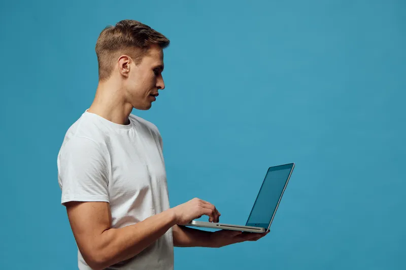Young man in casual white t-shirt using a laptop computer with a blue background, standing in profile, focused on work or browsing, modern technology workspace, people lifestyle concept, isolated on b