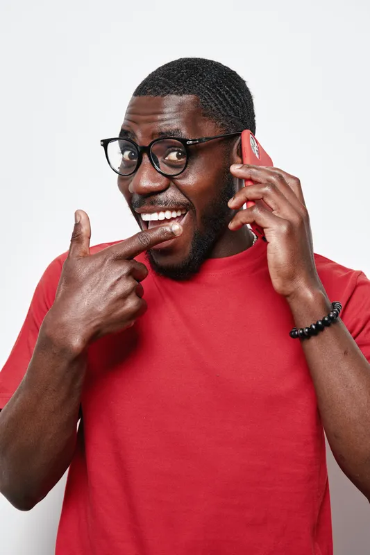 African American man with dark skin wearing glasses and red shirt talking on smartphone smiling and pointing finger at mouth isolated on white background with happy expression.