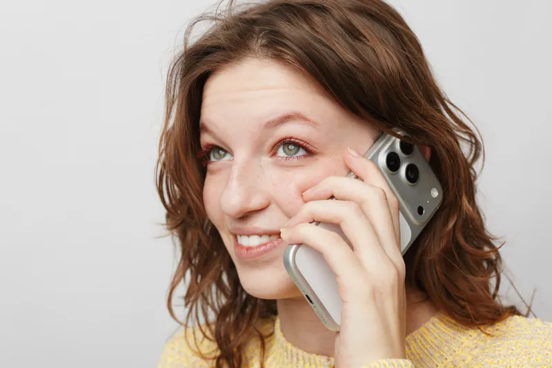 Young woman with curly hair smiling while talking on smartphone. Close-up portrait on white background. Concept of communication, technology, and positive emotions.