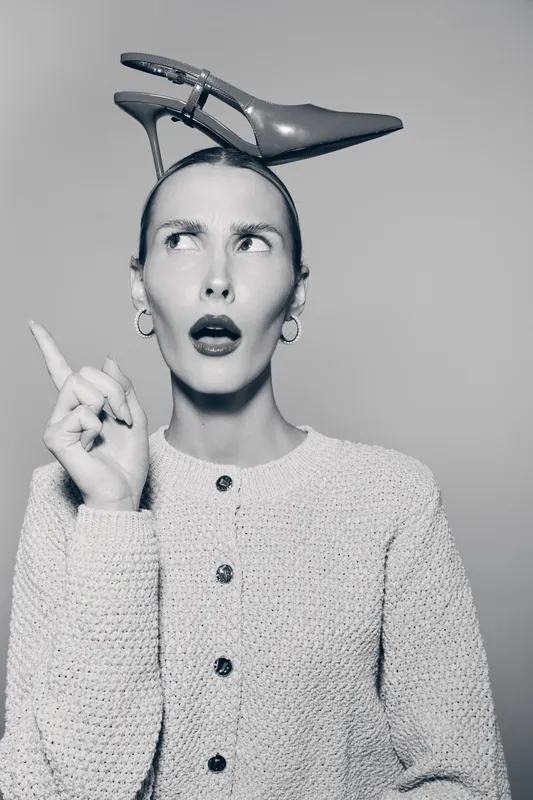 Confused white woman with pale skin and earrings wears high heel shoe on head and points up. Concept of fashion and unusual style.