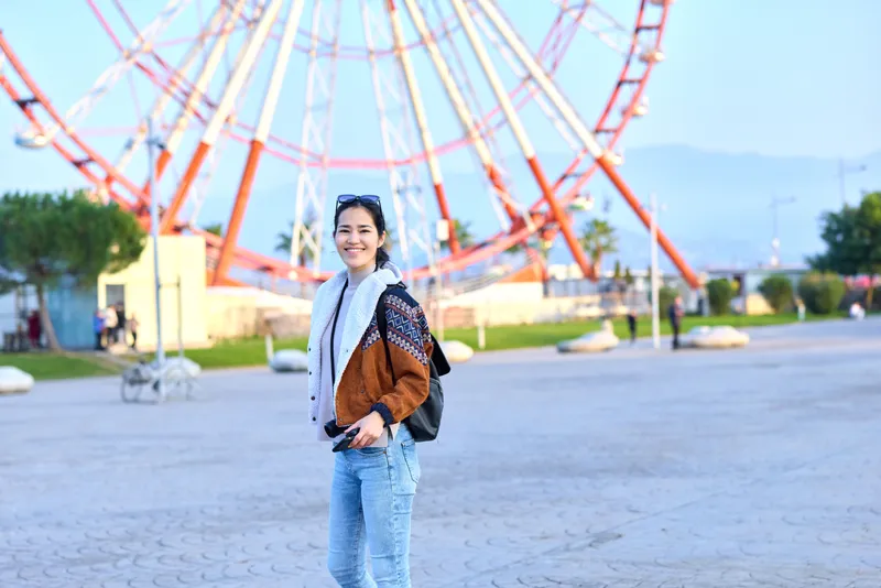 Woman traveler in a casual jacket and scarf stands in an outdoor park beside a ferris wheel, smiling with a backpack, capturing sunny amusement and urban leisure.