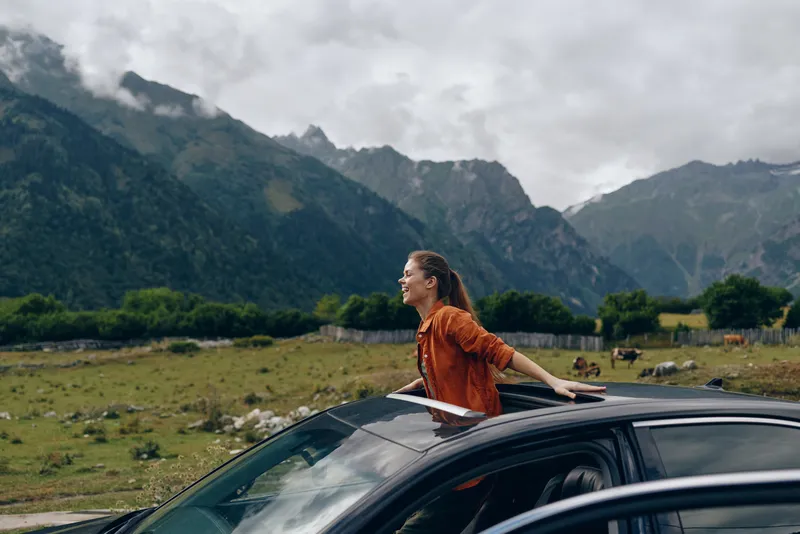 person car mountains travel roadtrip adventure freedom inclusivity, woman leaning from car sunroof in rural field, authentic inclusive portrait joyful outdoors under cloudy sky