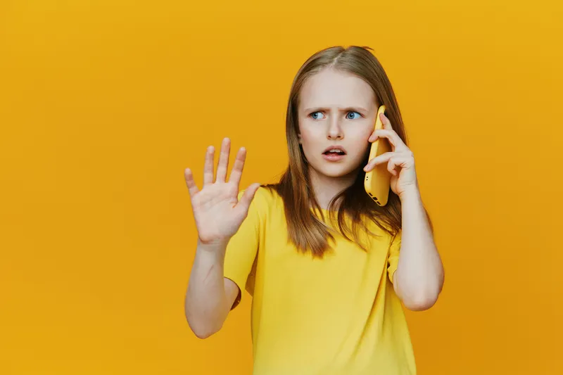 Young girl expressing surprise while talking on the phone against a bright yellow background, showcasing a casual style and emotional response