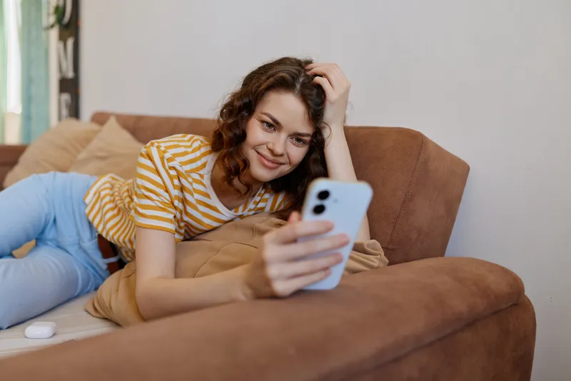 Young woman lounges on a cozy sofa at home, casually scrolling on her smartphone with a light, amused smile, as soft natural light filters through the window, creating an authentic, relatable