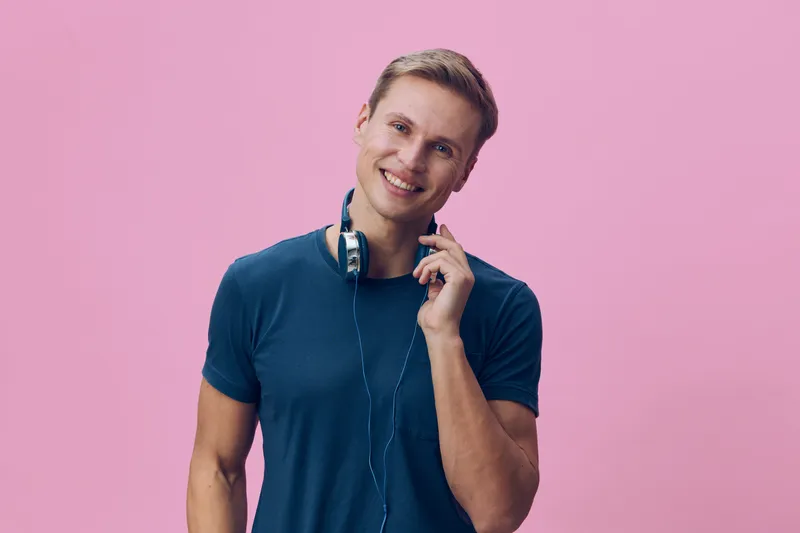 Smiling young man wearing casual blue t-shirt holding earphones in hand, looking cheerful against pink background. Studio portrait of happy man listening to music, casual style, positive mood, lifesty
