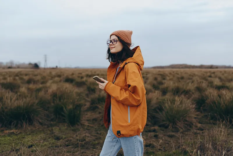 Woman lifestyle outdoors in autumn field, nature grassland, wearing orange jacket, beanie and glasses, with smile while walking, casual travel mood, soft daylight, breezy