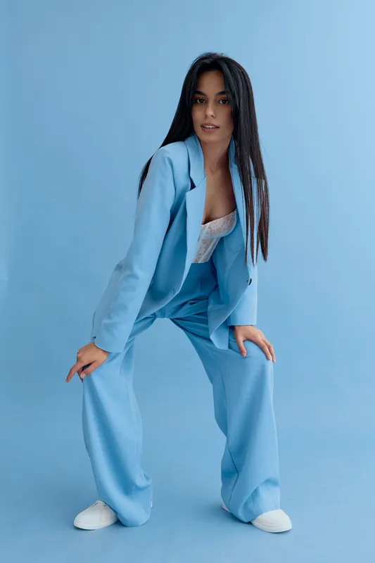 Stylish woman wearing a loose sky-blue suit and white top posing against a blue studio background with long dark hair and casual white sneakers, modern fashion style.