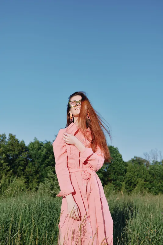 Young woman in pink dress enjoying nature, with long hair blowing in the wind against a sunny blue sky and green background