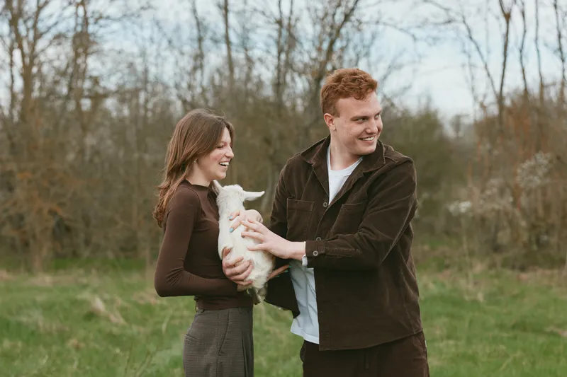 Young couple enjoys holding a small white baby goat outdoors in a grassy field during autumn, sharing a joyful moment in casual warm clothing.