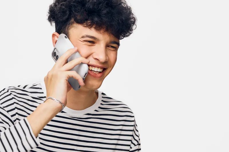 Young man smiling while talking on smartphone, wearing a striped shirt with curly hair and joyful expression on white background, communicating happily.
