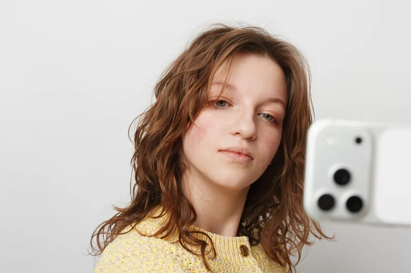 Young woman with curly hair wearing yellow sweater takes selfie with smartphone. Casual style, natural light, and neutral background create relaxed and authentic portrait concept.