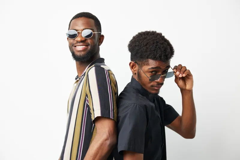 Two stylish African American men smiling together, wearing sunglasses and fashionable shirts, exuding confidence and friendship while posing against a clean white background.