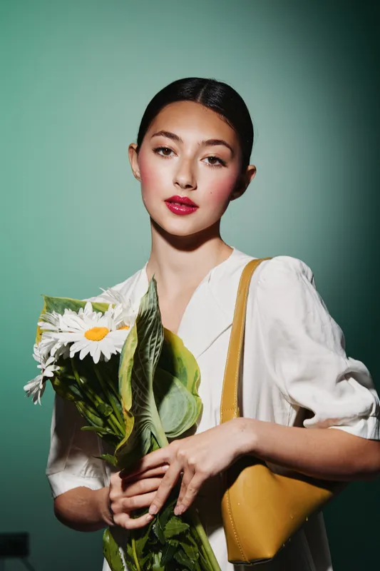 Young woman in a white blouse holds daisies and green leaves, carrying a yellow bag over her shoulder. Beauty, fashion, and creative floral concept with soft studio green background.