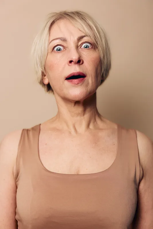 Surprised middle aged woman with short blonde hair stares wide eyed and open mouthed at camera, wearing beige sleeveless top against neutral background. Concept for shock, amazement, emotion.