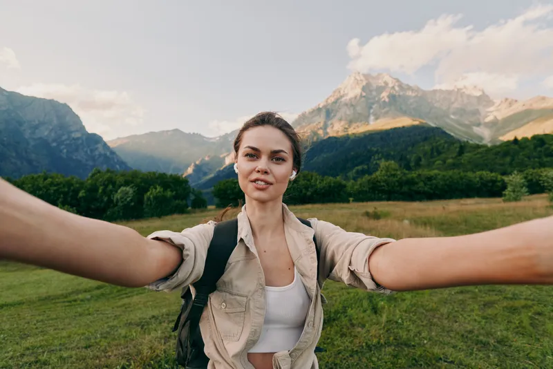 Young woman with brunette hair takes a selfie while hiking in a mountain valley during a sunny day, showing confidence and adventure spirit.