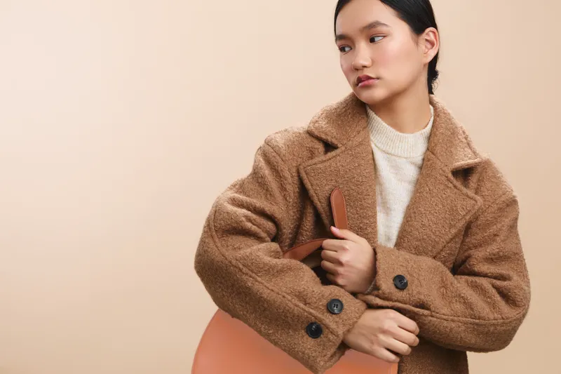 Asian woman stands in a warm coat against a neutral backdrop, posing with calm confidence and thoughtful gaze, conveying a poised, stylish mood and contemporary vibe. Her posture and subtle smile