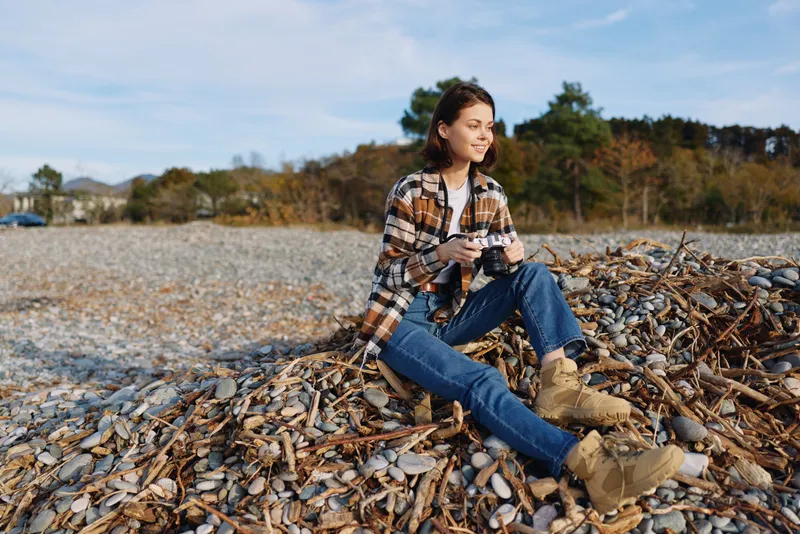 Woman outdoors with camera sits on a leaf pile near a pebble beach, autumn light, casual plaid shirt, blue jeans and boots, calm creative mood for nature photography