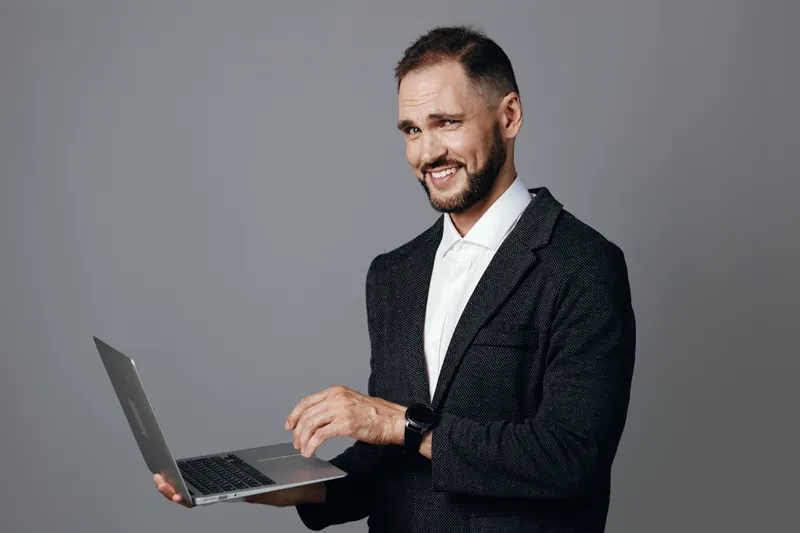 A professional businessman stands with a laptop against an isolated gray background, projecting confidence, focus, and calm optimism during digital work.