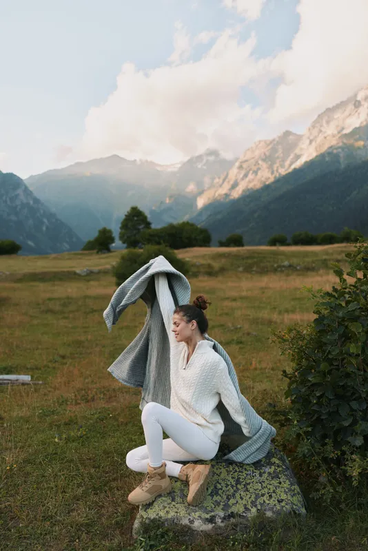 Young woman wearing white sweater and boots sits on a mossy rock in a wide meadow with dramatic mountains in the background, wrapped in a gray blanket for a calm outdoor moment.